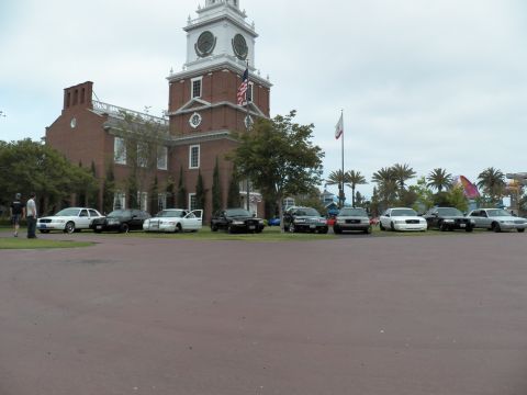 2003 Ford Crown Victoria - Black and white sedans line the grass in front of a brick clock-tower hall and palm trees.