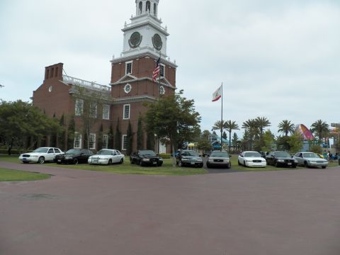 2003 Ford Crown Victoria - Crown Victoria and Grand Marquis sedans line a grassy lot beside a brick clock tower under overcast skies.