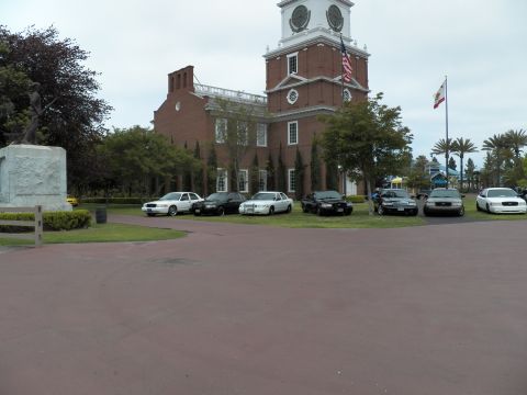 2003 Ford Crown Victoria - Ford and Mercury sedans sit on grass beside a bronze statue and the brick clock-tower facade.