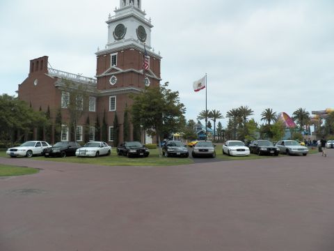 2003 Ford Crown Victoria - Ford sedans gather on lawn with amusement-park rides and palms visible past the flagpoles.
