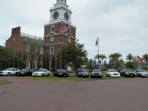 2003 Ford Crown Victoria - Ford and Mercury sedans line the grass below the clock tower and twin flagpoles.