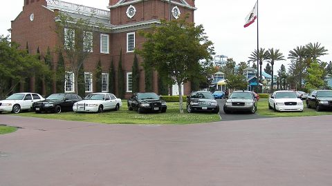 2003 Ford Crown Victoria - Ford sedans line the lawn with palms and a California flag behind the brick facade.