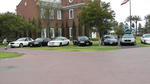 2003 Ford Crown Victoria - Panther-platform sedans sit on lawn near the brick clock-tower wall and shade trees.