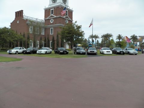 2003 Ford Crown Victoria - Ford and Mercury sedans gather on the lawn under flags with theme-park rides beyond.