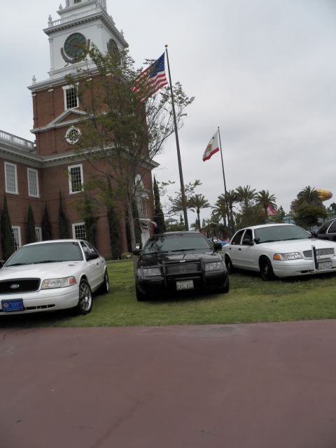 2003 Ford Crown Victoria - White, black and white Crown Victorias sit on lawn below the flags and clock tower.