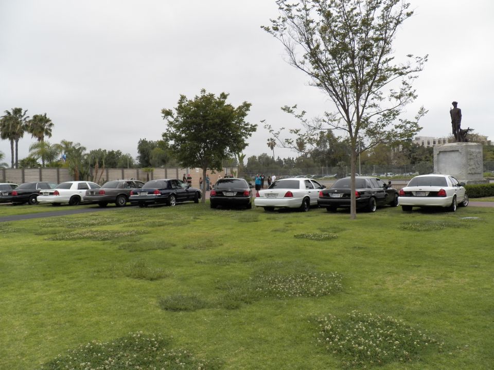 Row of full-size sedans parked on grass near a bronze statue and brick building