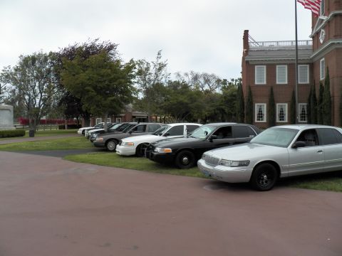 2003 Ford Crown Victoria - Silver sedan fronting a lineup of Ford and Mercury cars beside a brick building with flag
