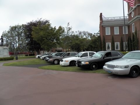2003 Ford Crown Victoria - Silver sedan with chrome grille at the end of a lineup near a flagpole and brick hall