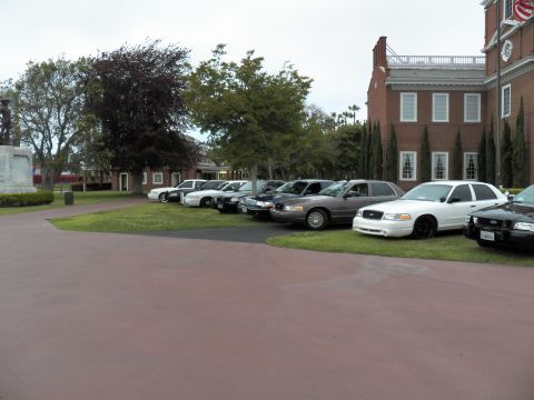2003 Ford Crown Victoria - White, silver and black sedans lined on a lawn with tall cypress trees and brick hall behind