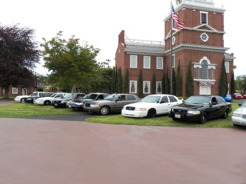 2003 Ford Crown Victoria - Lineup of Crown Victoria and Mercury sedans by a brick building with bunting and flag