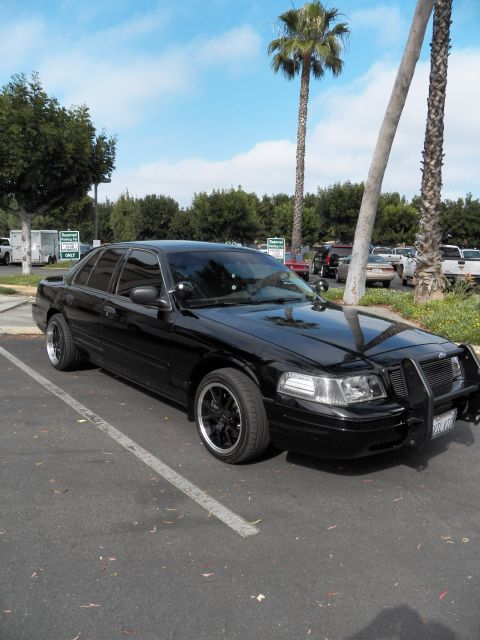 2003 Ford Crown Victoria - Murdered-out Crown Victoria parked front-three-quarter, palm trees behind, brush guard up front.