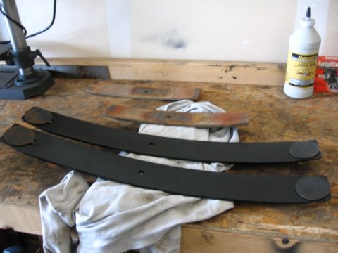 1995 Ford Bronco - Two new black leaf springs and two old rusty leaf springs are laid out on a workbench.
