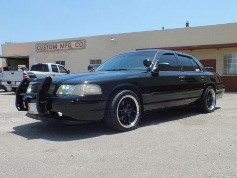 2003 Ford Crown Victoria - Front-three-quarter of murdered-out Crown Vic with brush guard, white truck beside.