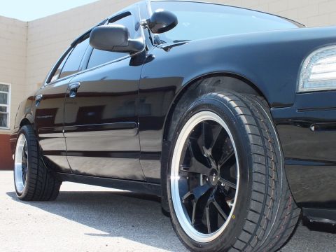 2003 Ford Crown Victoria - Front three-quarter of a lowered black Ford sedan showing the door, mirror and black polished-lip wheel.