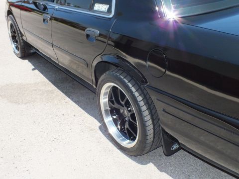 2003 Ford Crown Victoria - Low side view of a black Ford sedan's rear wheel, fuel door and Ford-logo mud flap.