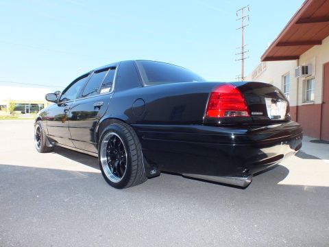 2003 Ford Crown Victoria - Rear quarter of a glossy black Crown Victoria against a clear sky, riding low on staggered black wheels.