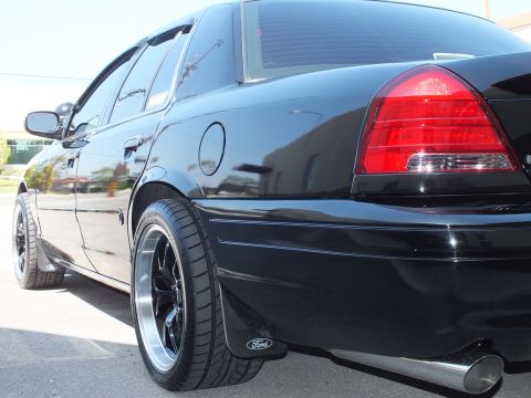 2003 Ford Crown Victoria - Glossy black Crown Victoria rear quarter shot low to the ground, chrome exhaust tip visible beneath bumper.