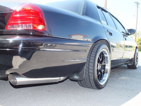 2003 Ford Crown Victoria - Ground-level black Crown Victoria rear quarter, chrome exhaust tip and Ford mud flap above black wheel.