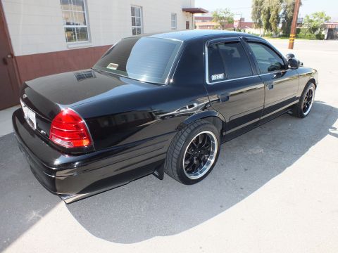 2003 Ford Crown Victoria - Rear three-quarter of a glossy black Crown Victoria parked by a wall, low stance on black machined wheels.