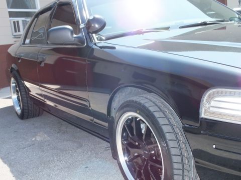 2003 Ford Crown Victoria - Front-quarter close-up of a black Crown Victoria, headlight cluster and black wheel under bright sun flare.