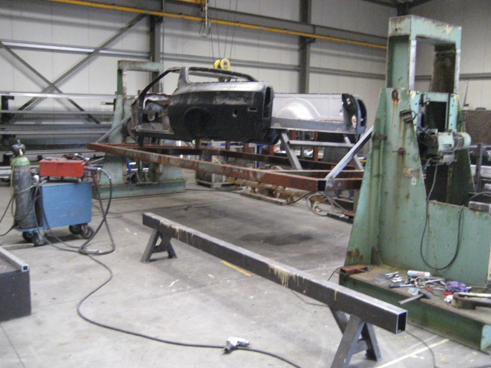Car body suspended on a metal frame in a workshop, with welding equipment nearby