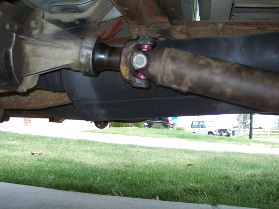 Underneath view of a driveshaft connected to a differential, with a fuel tank visible. Two trucks are parked in the background