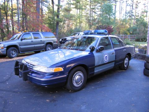 2001 Ford Crown Victoria - A two-tone police car is parked on a driveway with a suburban and trees in the background.