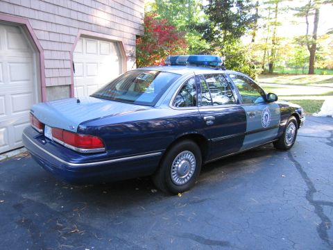 2001 Ford Crown Victoria - A two-tone blue police car with a light bar is parked in front of a garage.