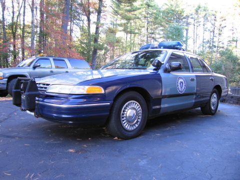2001 Ford Crown Victoria - A police car with a push bumper and light bar is parked outside next to a pickup truck.