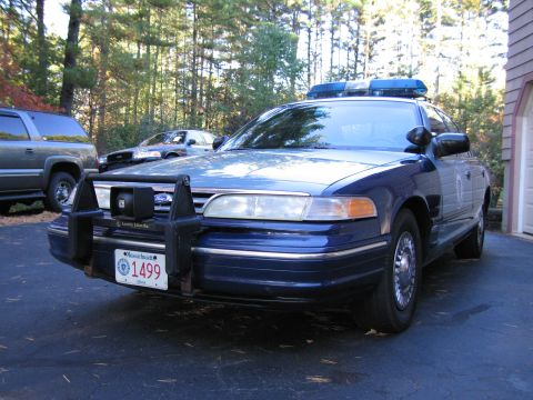 2001 Ford Crown Victoria - A blue 2001 Ford Crown Victoria police car is parked in front of a garage, with other vehicles visible in the background.