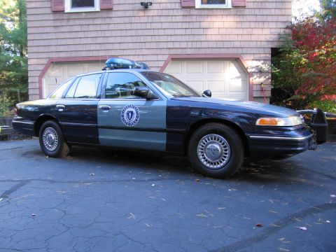 2001 Ford Crown Victoria - A two-tone police car with a light bar is parked in front of a garage.