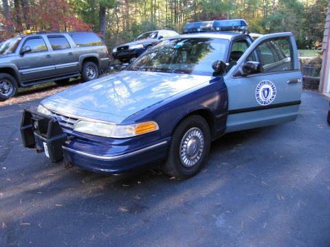 2001 Ford Crown Victoria - A Massachusetts State Police car, a 2001 Ford Crown Victoria, is parked with its door open, showing a police emblem on the side.