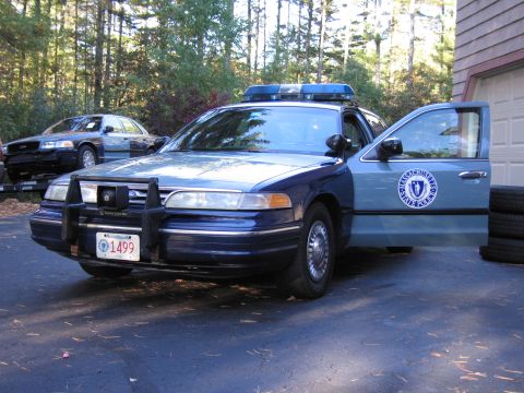 2001 Ford Crown Victoria - A Massachusetts State Police car is parked in a driveway with its door open, and another police car is visible in the background.