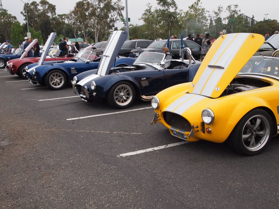 Yellow Shelby Cobra replica with hood open, parked in a lot with other similar cars