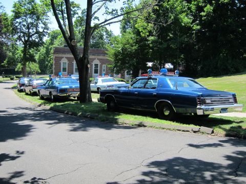 1991 Ford LTD Crown Victoria - line of classic police cruisers parked beside a grassy lawn, with a blue State Police sedan closest to the camera.