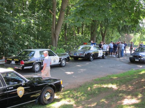 1991 Ford LTD Crown Victoria - Spectators mingle among several parked vintage police cars on a tree-shaded street at a car show.