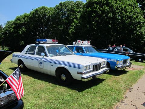 1991 Ford LTD Crown Victoria - white Police sedan with blue stripes parked beside a blue Police car on a sunlit lawn, a small US flag nearby.