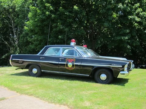 1991 Ford LTD Crown Victoria - black vintage police sedan with a shield-shaped door emblem and a single red rooftop beacon, parked on a lawn.