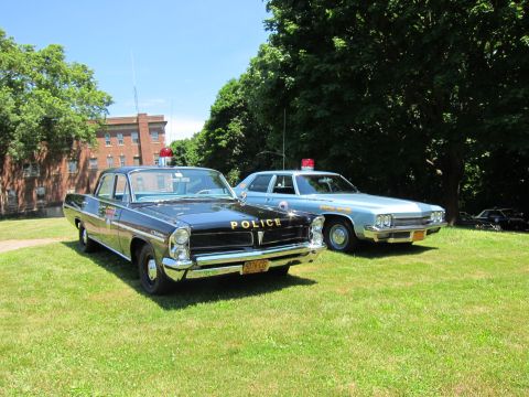 1991 Ford LTD Crown Victoria - black Police-marked sedan with a red rooftop beacon parked beside a light blue cruiser on a lawn.