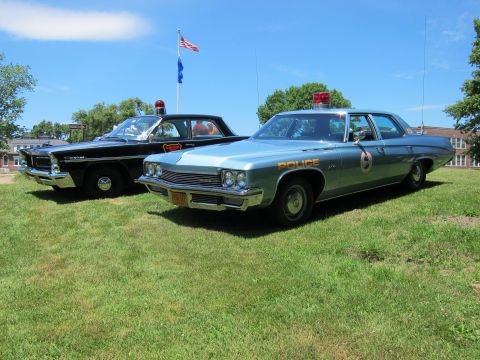 1991 Ford LTD Crown Victoria - light blue Police-marked sedan parked beside a black cruiser on a lawn with an American flag in the background.