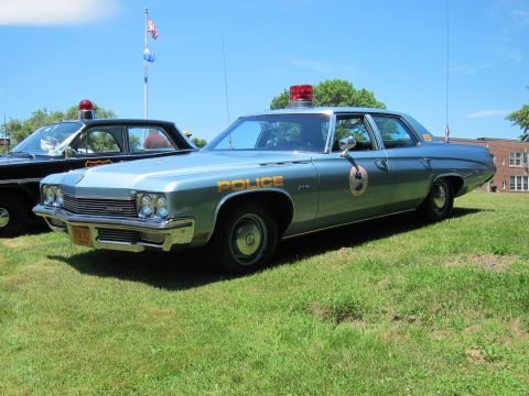 1991 Ford LTD Crown Victoria - silver-blue Police-marked sedan with a red rooftop beacon and shield door emblem parked on a lawn.