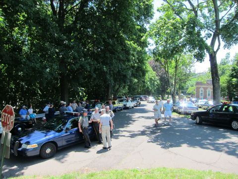 1991 Ford LTD Crown Victoria - crowd mingles among modern and vintage police cars on a sunny tree-lined street, with a STOP sign at left.