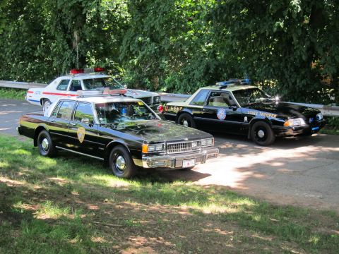 1991 Ford LTD Crown Victoria - Three police cars parked on a wooded drive, including a black sedan, a white cruiser, and a black State Trooper Mustang.