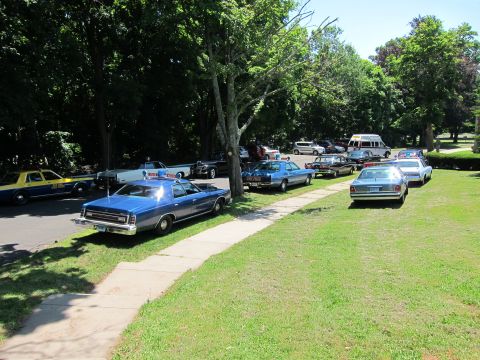 1991 Ford LTD Crown Victoria - Numerous vintage police cars parked along a tree-lined drive next to a concrete walkway and grassy lawn.