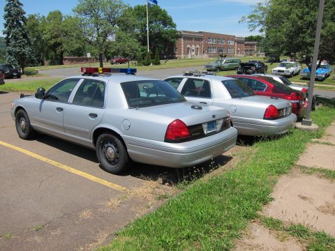 1991 Ford LTD Crown Victoria - Two silver police cruisers with light bars parked nose-to-curb in a lot, with a brick school building behind.