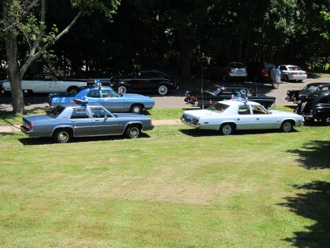 1991 Ford LTD Crown Victoria - Two light-blue classic police sedans with roof lights parked on a grassy field among other show vehicles.