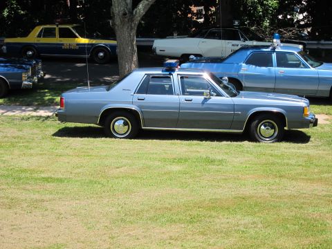 1991 Ford LTD Crown Victoria - light-blue classic police sedan with a roof light shown in side profile on a grassy field, other cruisers behind.