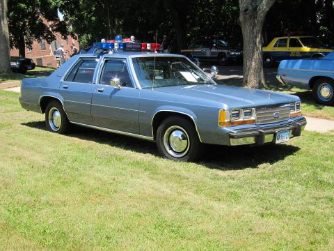 1991 Ford LTD Crown Victoria - light-blue Ford police sedan with roof light bar and front spotlight parked on grass at a show.
