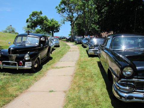 1991 Ford LTD Crown Victoria - Black classic 1940s-50s automobiles parked along both sides of a concrete walkway at a car show.