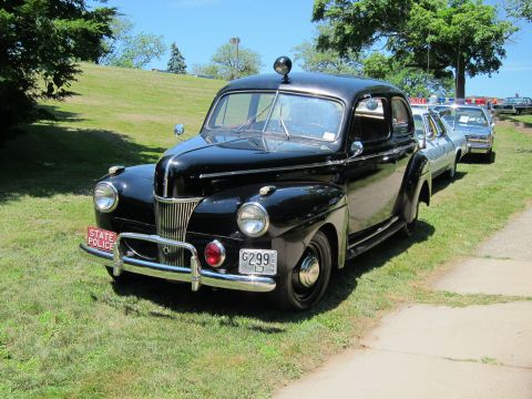 1991 Ford LTD Crown Victoria - black late-1940s Ford State Police sedan with red front plate parked on a grassy slope, cruisers lined behind.